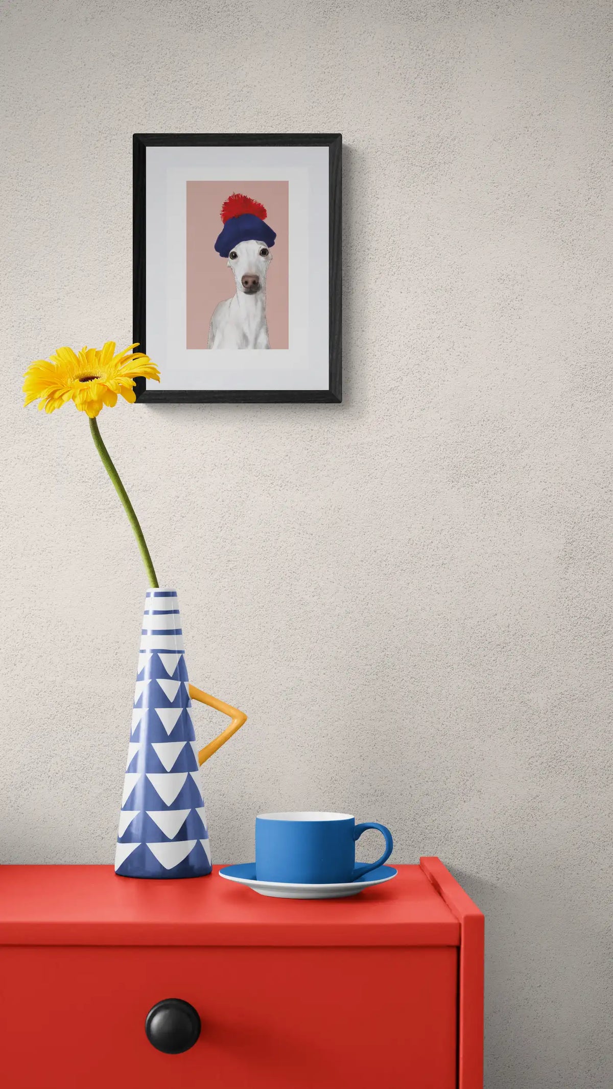 Kunstdruck des Motivs Iggy Souverän, weißer Iggy mit rot blauer Bommelmütze, in Din A4 auf Enviro Value Umweltpapier, gerahmt über roter Kommode mit Blumenvase und Tasse